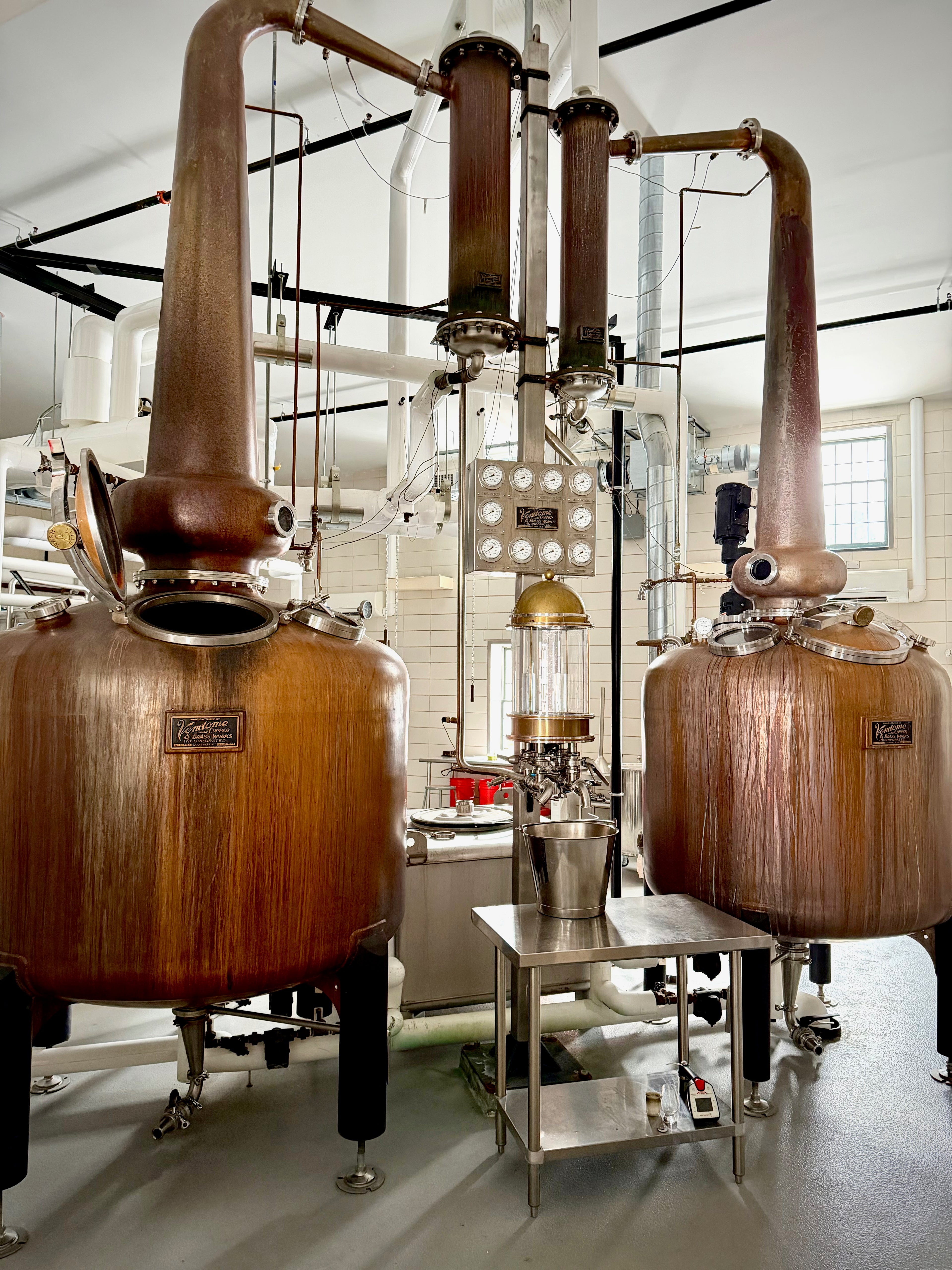 Large copper stills in a distillery setting with glass windows.