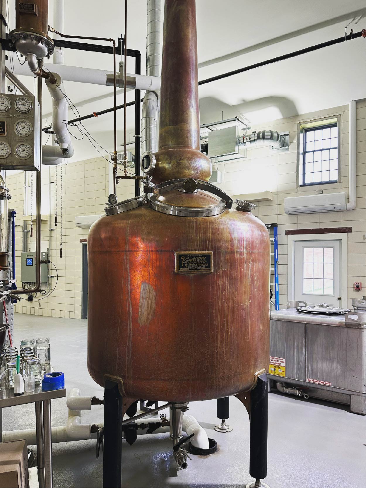 Copper still in a distillery setting with pipes and equipment in the background