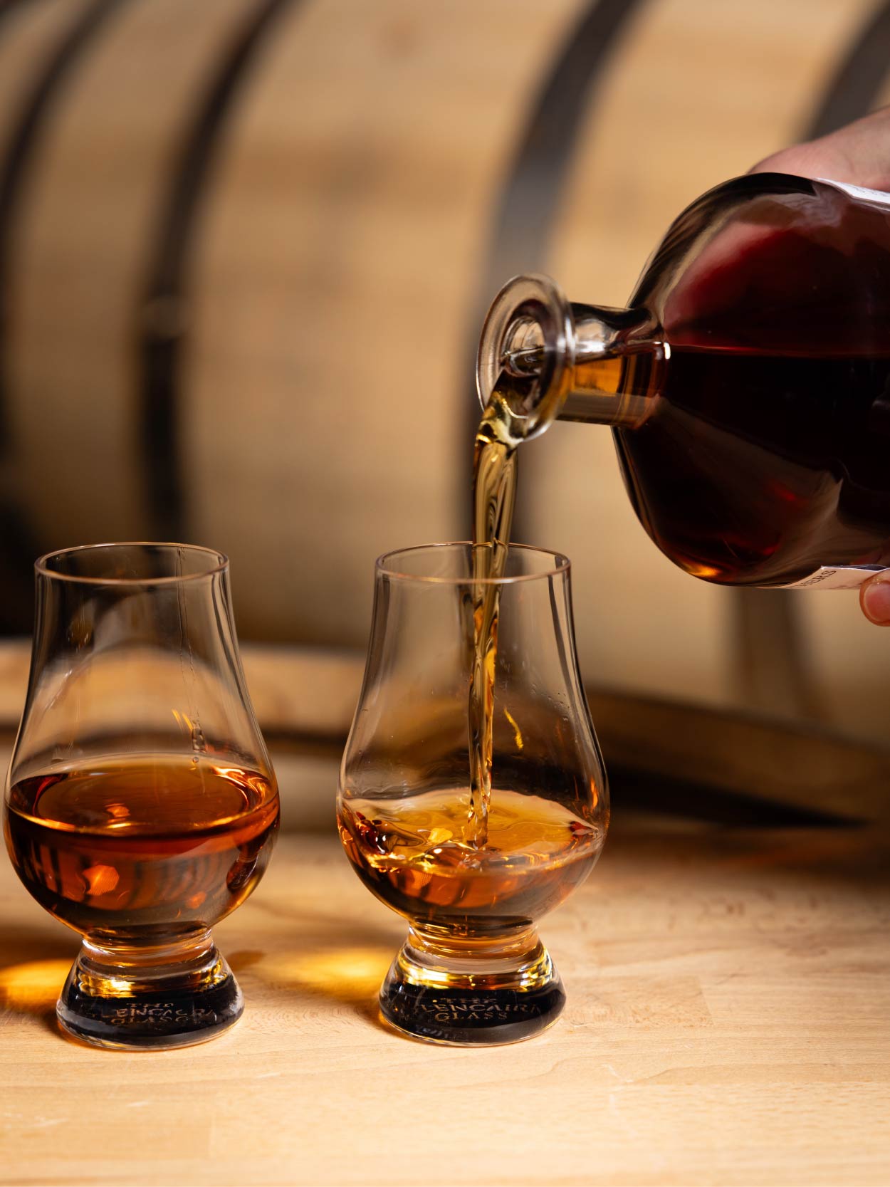 Whiskey being poured from a bottle into two glasses with a wooden barrel in the background