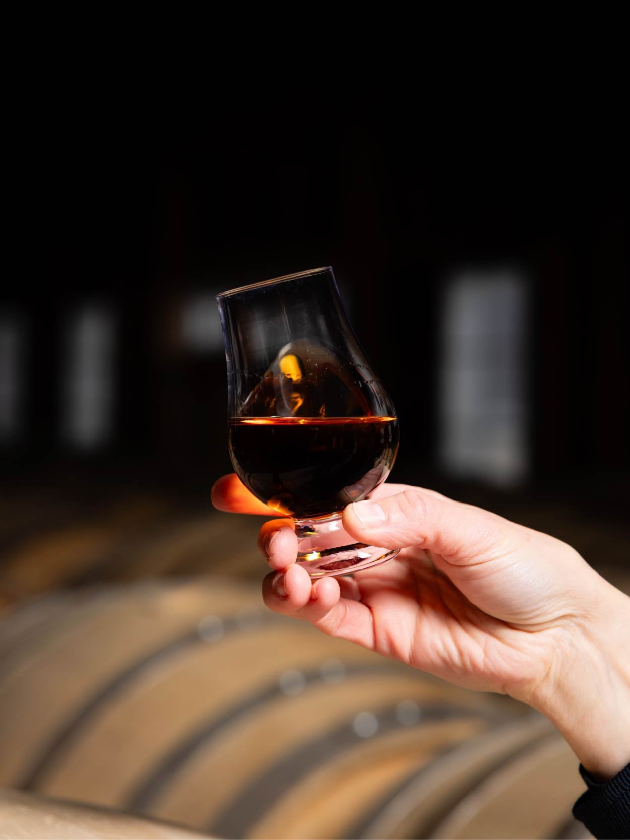 Hand holding a glass of whiskey in front of a blurred whisky barrel background