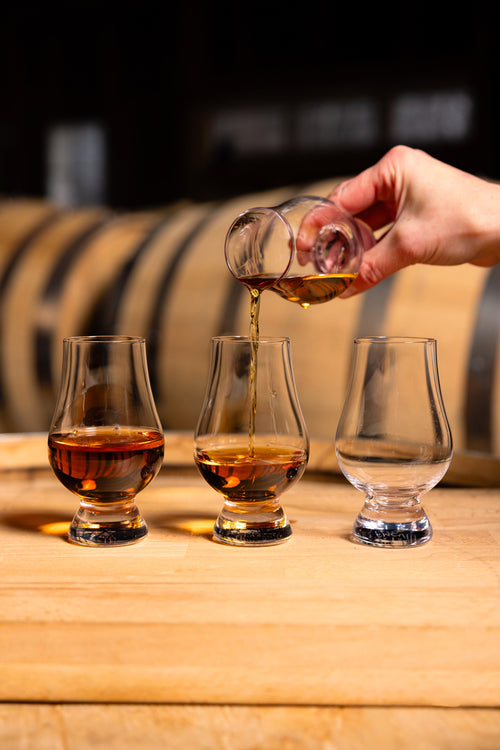 Hand pouring whiskey from one glass to another on a wooden surface with barrels in the background